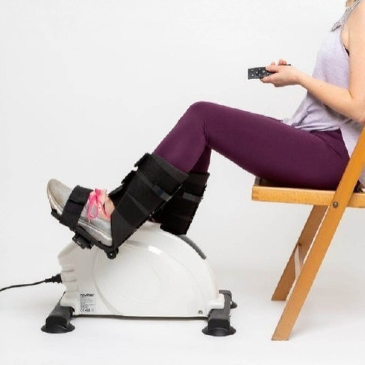 Seated on a wooden chair, a person uses the Reviber Mini Bike PRO by Reviber, feet strapped in and holding the remote. The motorised and manual mini exercise bike is plugged in, set against a plain white background.