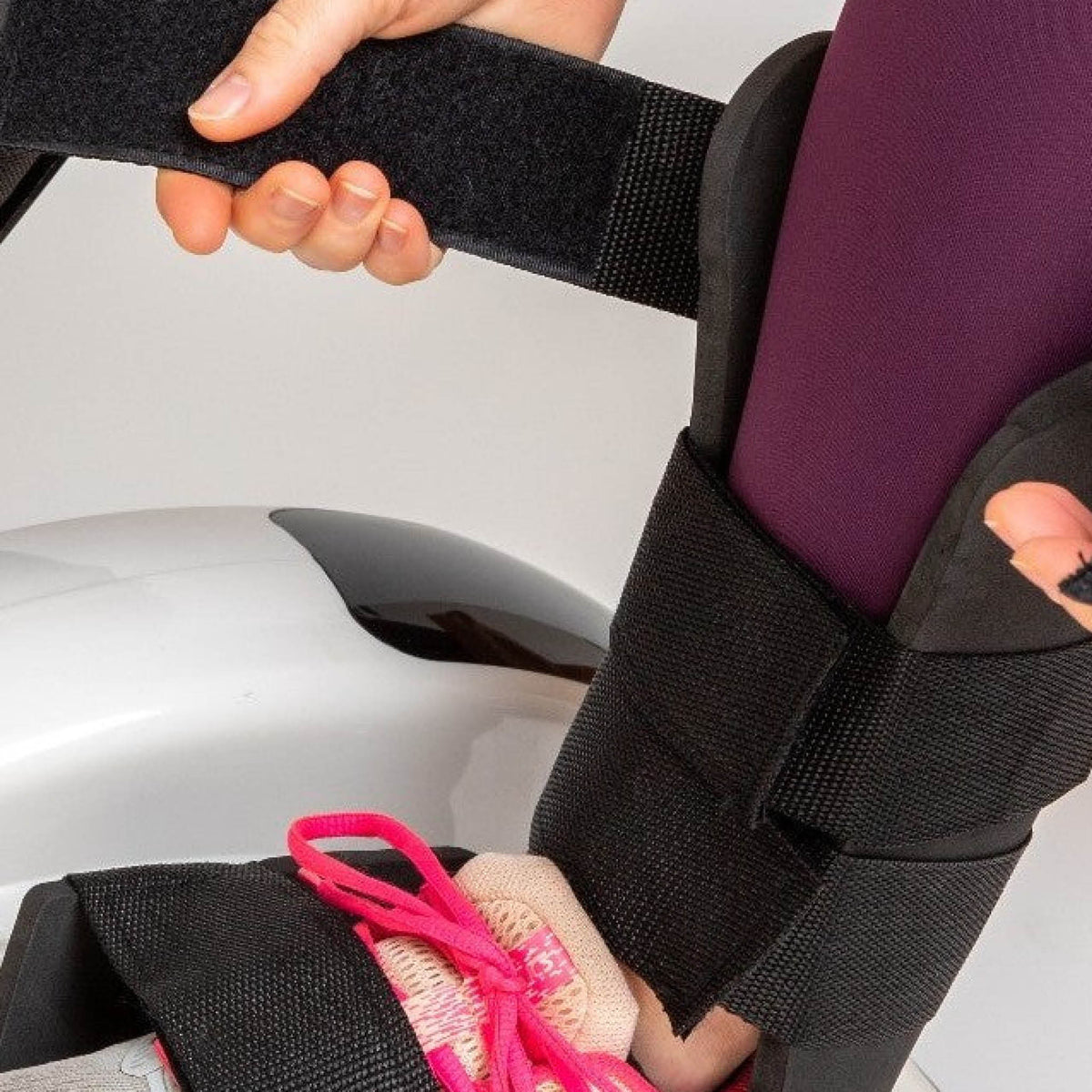 A person secures a black medical walking boot over their sneaker-clad foot and fastens the Velcro straps, preparing for low-impact rehabilitation using the Reviber Mini Bike PRO – a motorised and manual resistance exercise device from Reviber.