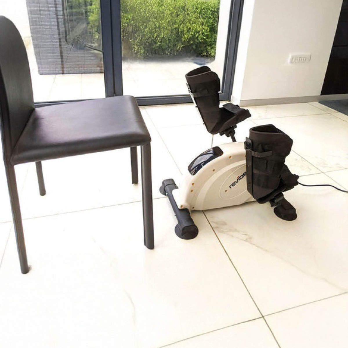 A chair faces the Reviber Mini Bike PRO, a motorised and manual resistance exercise machine with foot straps by Reviber, set on a tiled floor near large glass doors with green views—ideal for low-impact rehabilitation.
