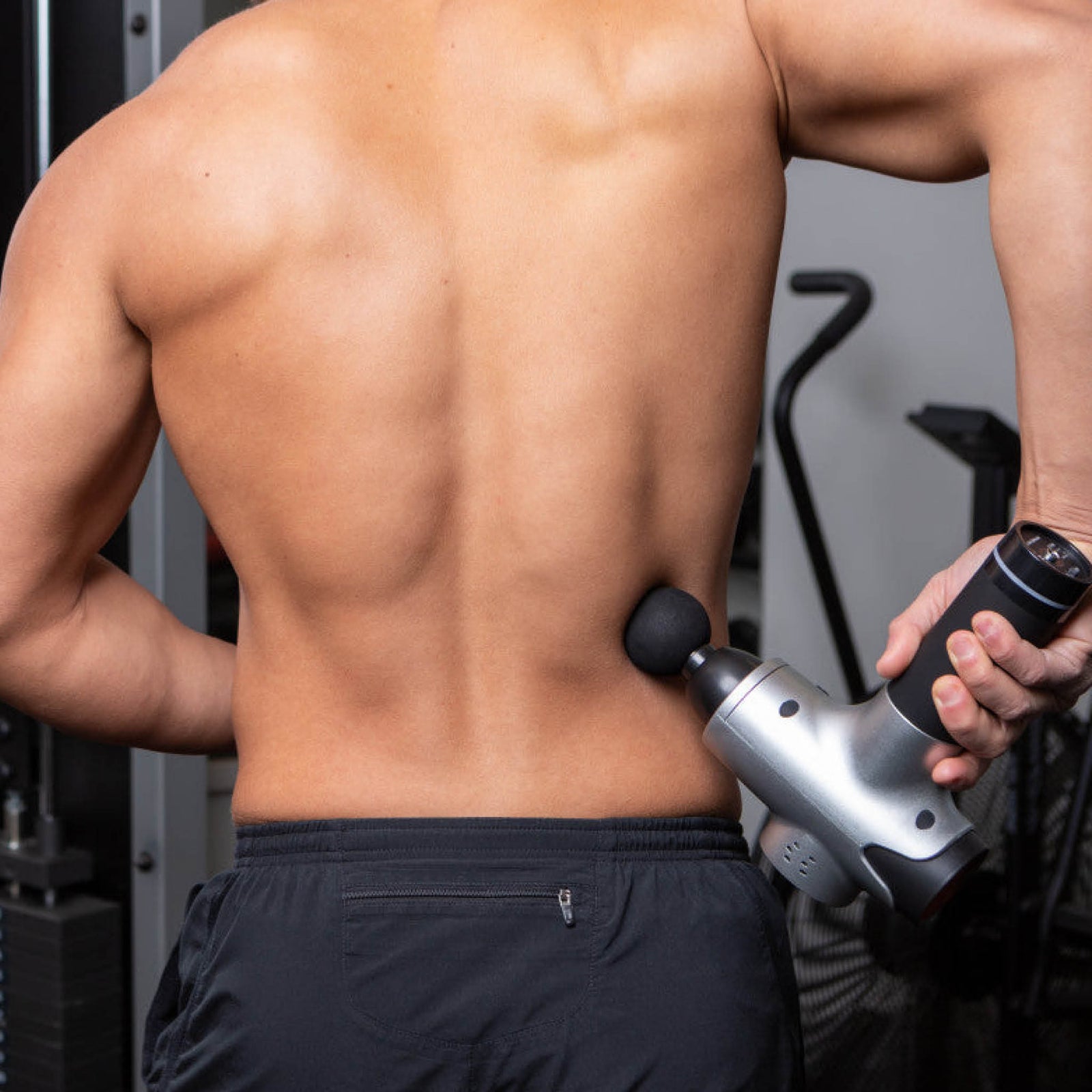 A shirtless person uses the Reviber Massage Gun by Reviber on their lower back in a gym, with exercise equipment visible in the background.