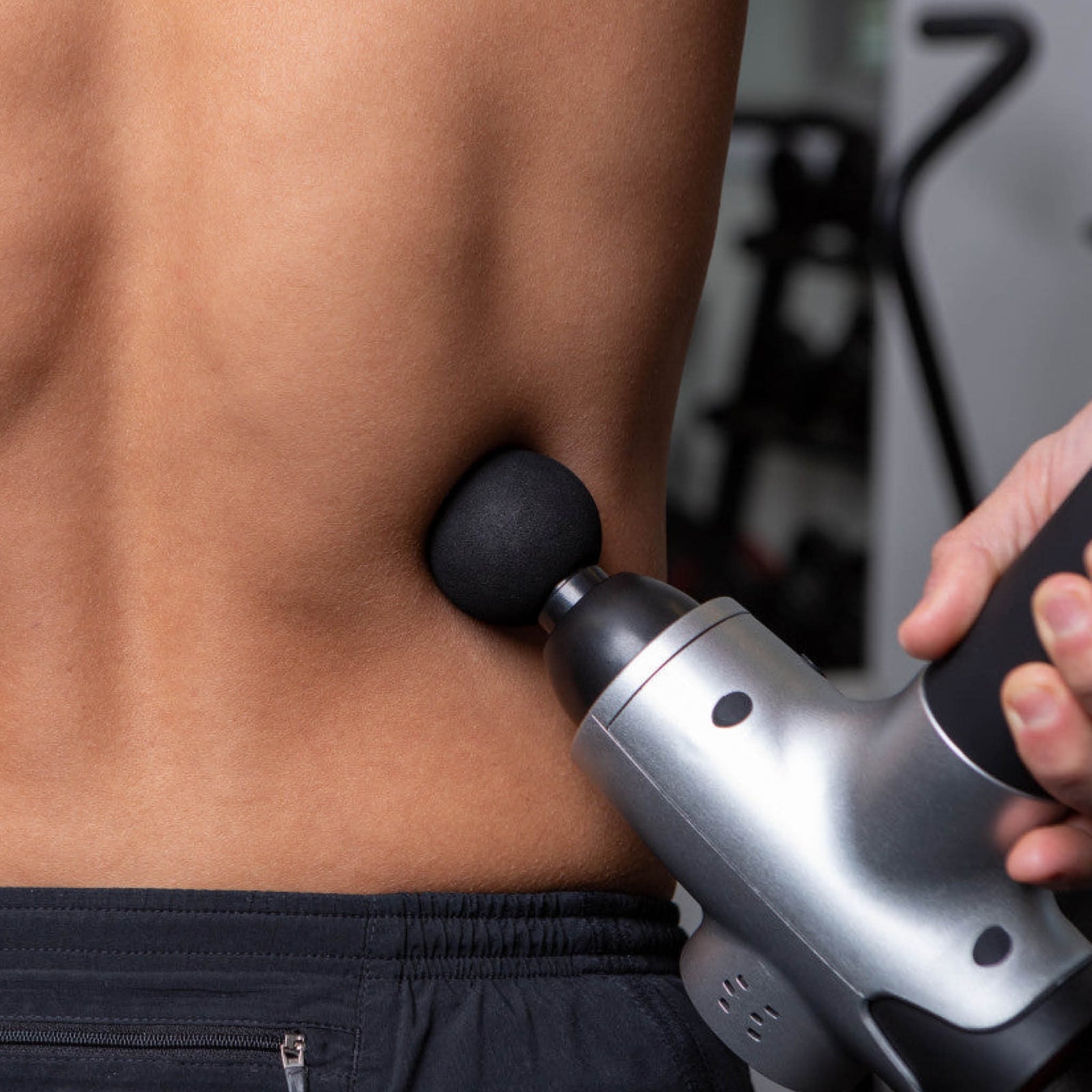 A person uses the Reviber Massage Gun with a round attachment on their lower back for deep-tissue massage, focusing on muscles. The blurred background highlights the Reviber device and the users bare skin.