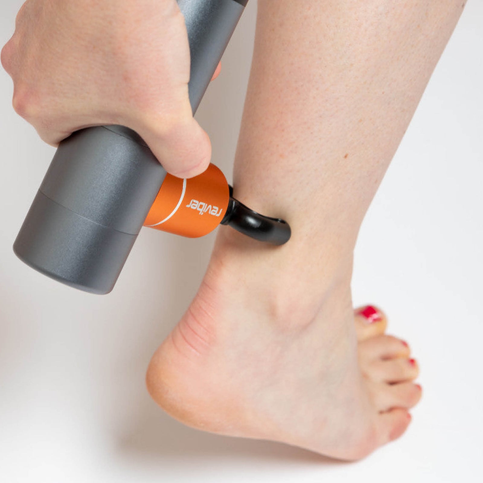A person uses the Reviber Mini Massage Gun With Heat Therapy by Reviber on their lower leg near the ankle. Their toenails are painted red, and a plain white background highlights this relaxing massage moment.